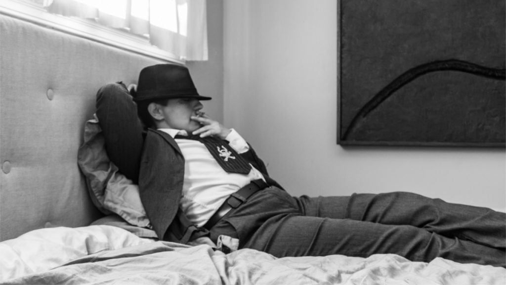 A black and white picture of a person in a suit and tie wearing a fedora low on their brow lounging on a bed propped up against the headboard, smoking a joint. 